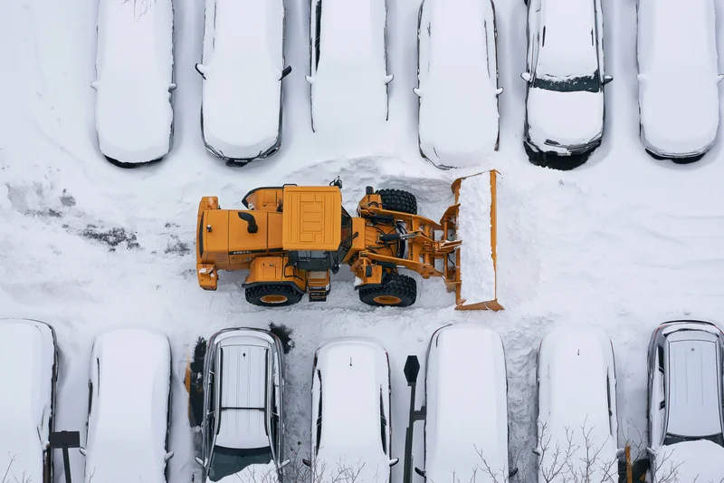 Yellow tractor clearing snow in a parking lot surrounded by snow-covered cars