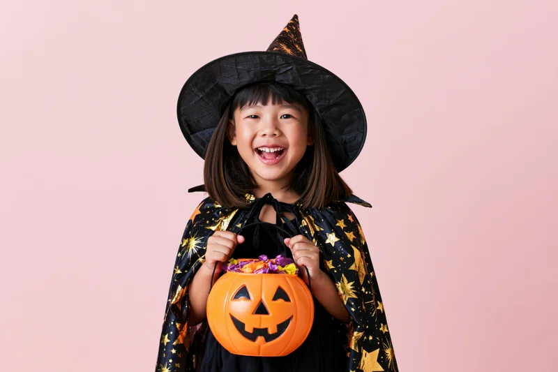 Young asian girl in witch costume holding pumpkin bucket of candy against pink background