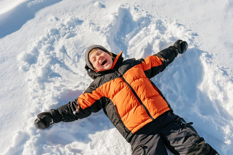 Young boy lying on snow making snow angel with arms and legs spread, laughing