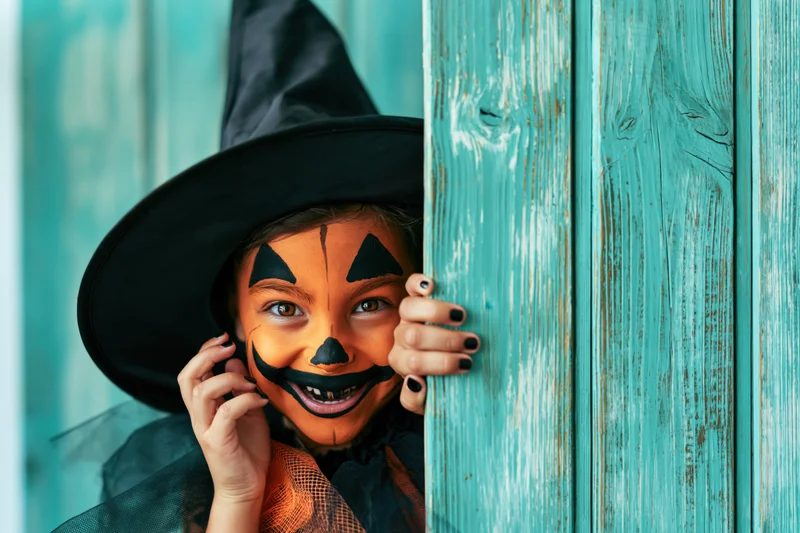 Child in witch costume with face paint peeking from behind teal wooden door