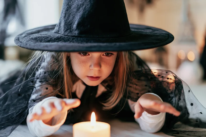 Young girl in witch costume leaning over candle with hands outstretched indoors