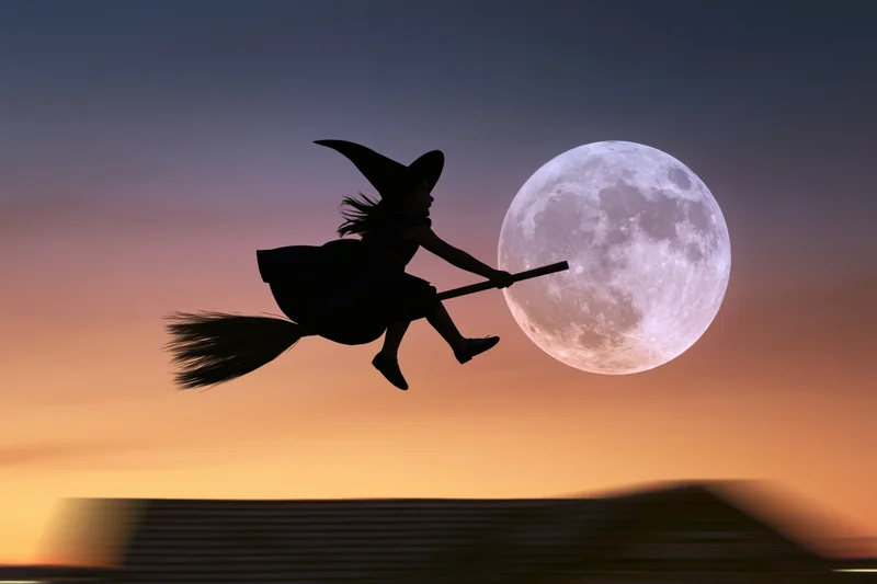Young girl in witch costume flying on broomstick silhouetted against full moon at twilight