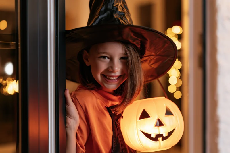 Young girl in witch costume smiling while peeking through a door holding a glowing jack-o-lantern lantern