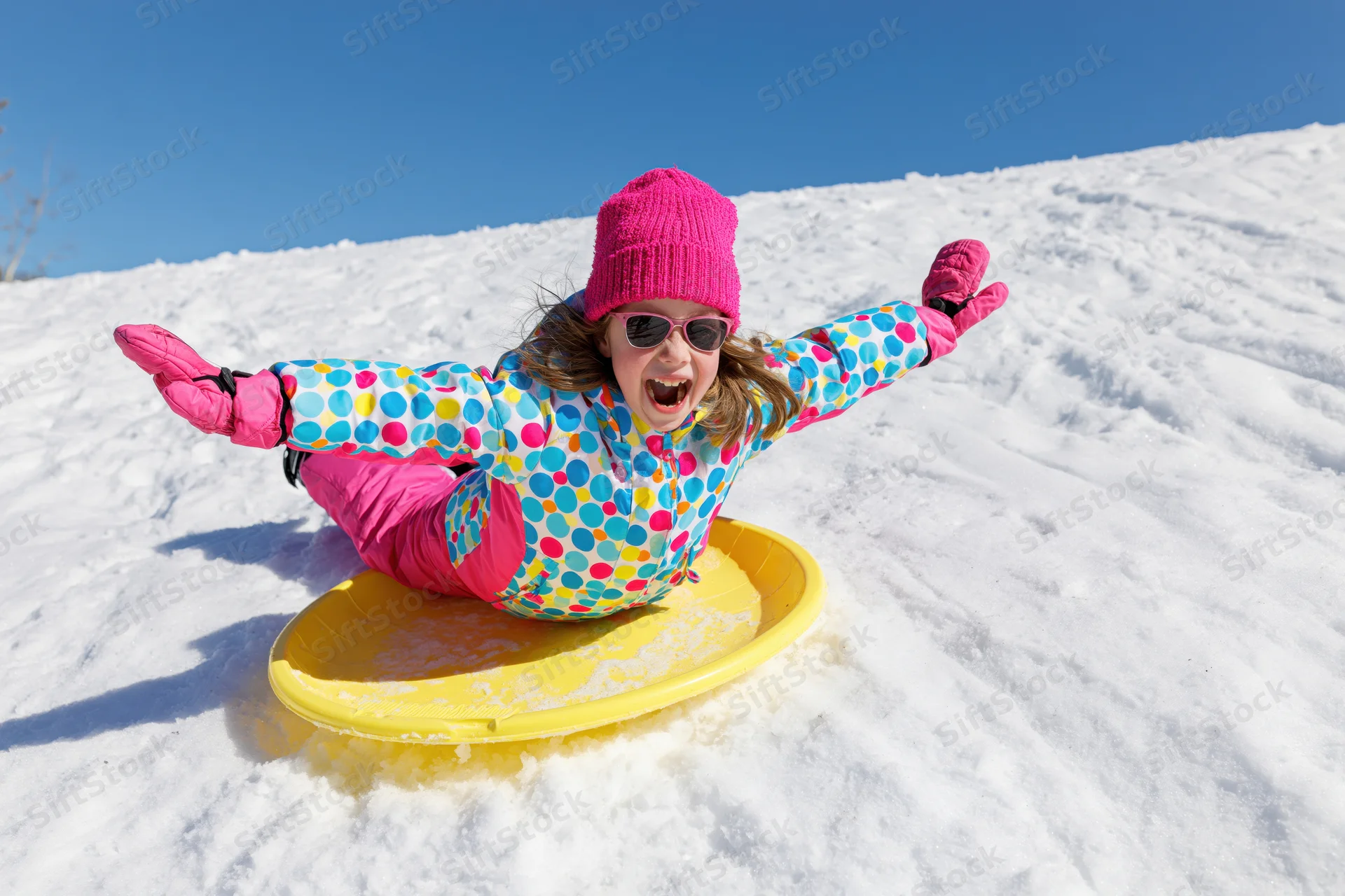 Young girl sledding down snowy hill on yellow plastic sled with arms outstretched and smiling
