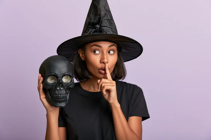 Young mixed-race woman in witch costume holding skull and making shushing gesture against purple background