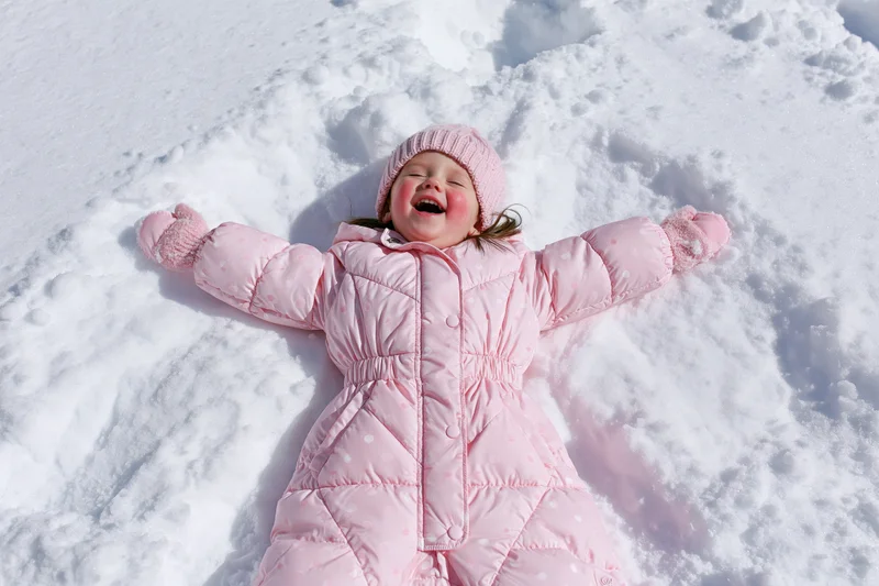 Toddler girl lying in snow making snow angel, laughing, wearing pink winter coat and hat
