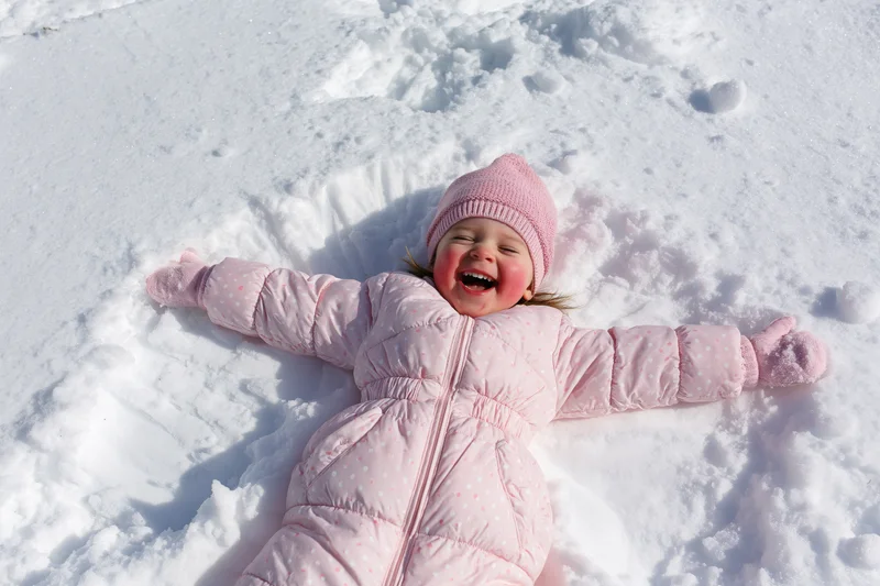 Toddler girl lying in snow making snow angel, laughing with arms and legs spread, wearing pink snowsuit and hat
