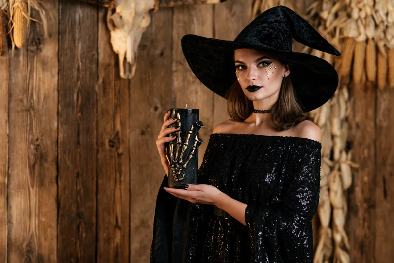 Woman in witch costume holding a dark candle against wooden wall with corn and skull decorations