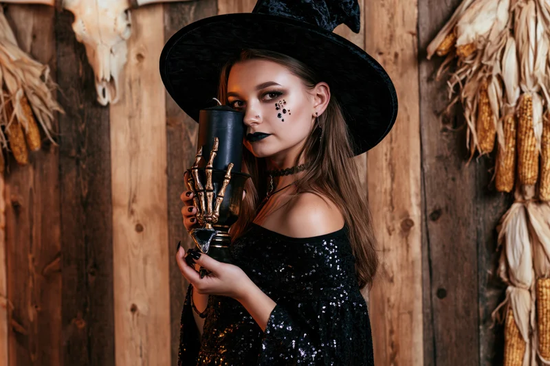 Woman in witch costume holding candle against wooden wall with corn and skull decorations