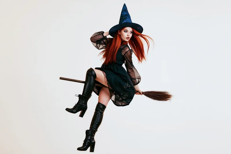 Young woman in witch costume riding broomstick against white background