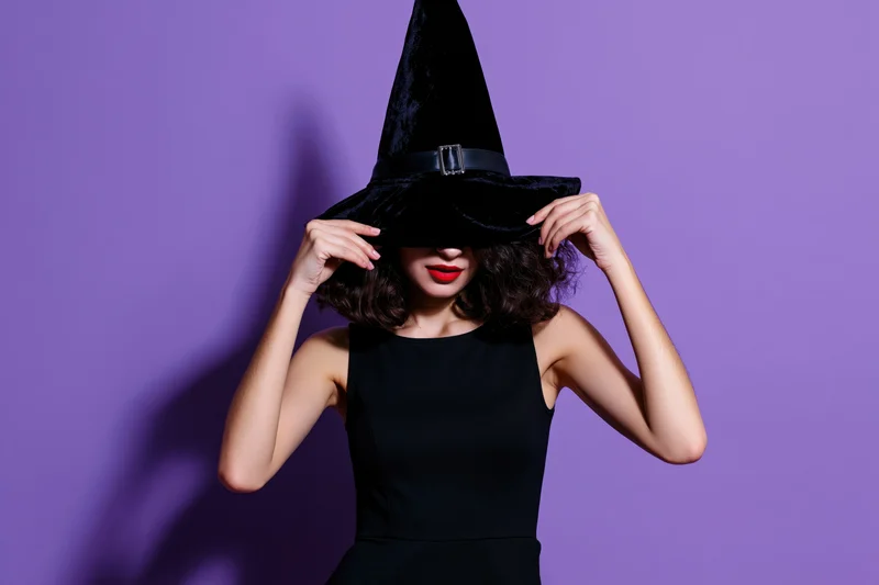 Woman in witch costume holding hat over face against purple background