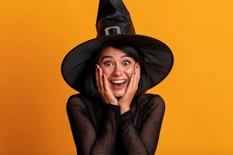 Woman in black witch costume smiling with hands on cheeks against orange background