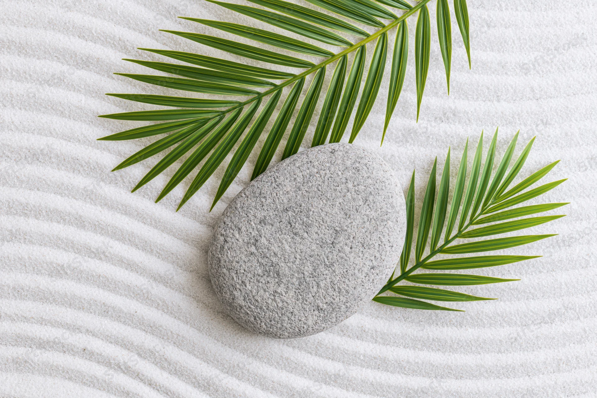 Smooth grey stone and green palm leaves arranged on raked white sand in a zen garden flat lay