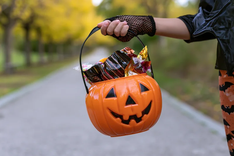 Child holding orange pumpkin-shaped candy bucket filled with sweets on a sidewalk lined with autumn trees