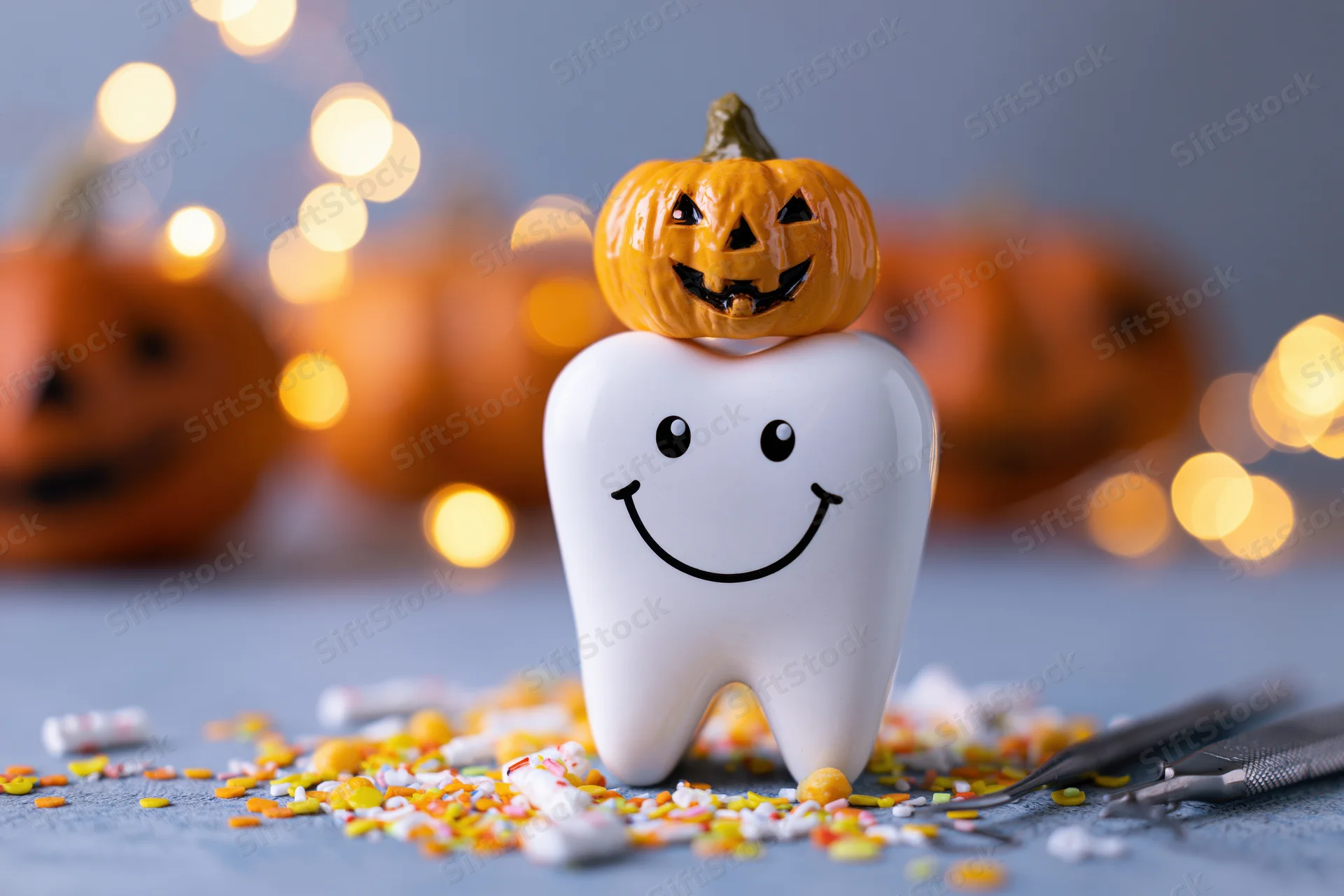 Ceramic tooth figurine with smiling face and small pumpkin on top surrounded by candy and dental tools on a gray surface with blurred pumpkins and lights in the background