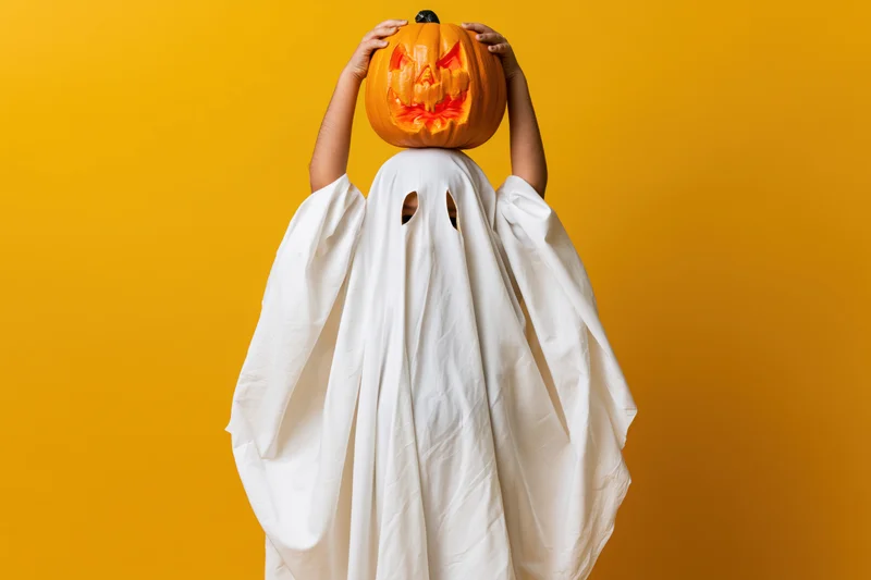 Child in white ghost costume holding carved pumpkin above head against yellow background