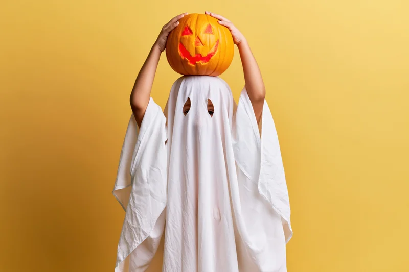Child in white ghost costume holding carved pumpkin above head against yellow background