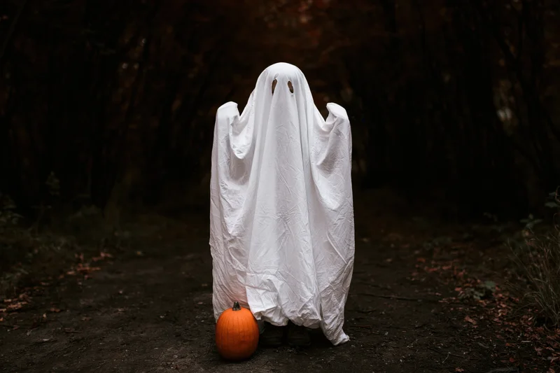 Child in white sheet ghost costume standing on forest path with pumpkin at feet