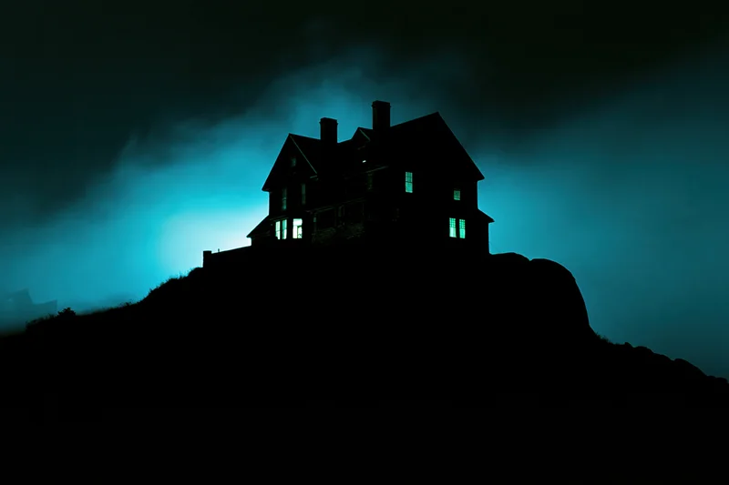 Dark silhouette of an old two-story house perched on a rocky hill at night with glowing windows and misty sky