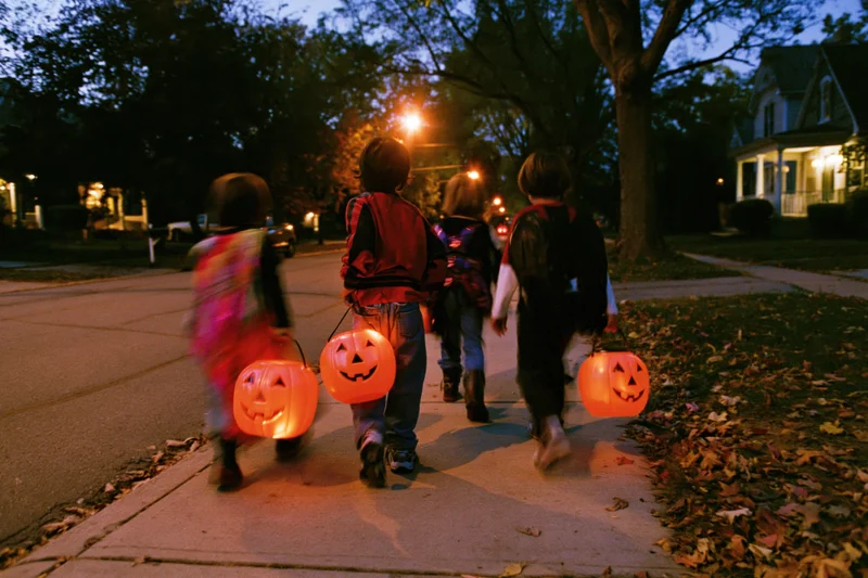 Children in halloween costumes walking down a suburban sidewalk at evening carrying glowing pumpkin lanterns