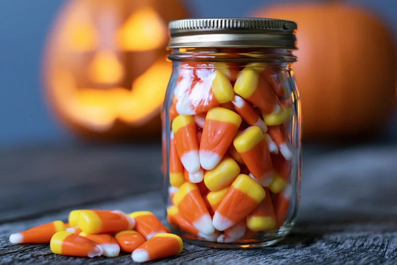 Candy corn in a glass mason jar on a wooden table with a glowing jack o lantern in the background