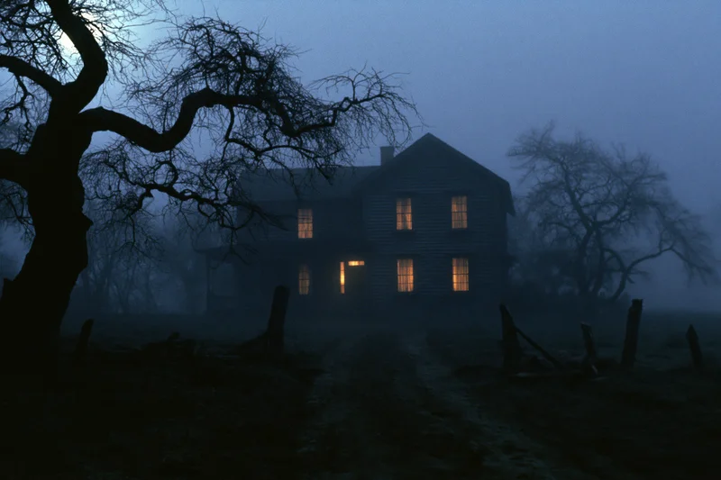 Isolated two-story wooden farmhouse with glowing amber windows at night in foggy rural landscape