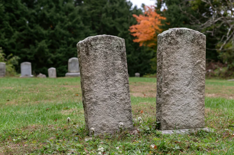 Two weathered granite gravestones standing in a grassy rural cemetery with autumn trees in the background