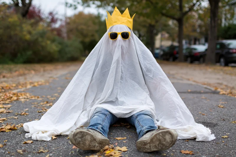 Child in white sheet ghost costume with yellow crown and sunglasses sitting on park path surrounded by autumn leaves