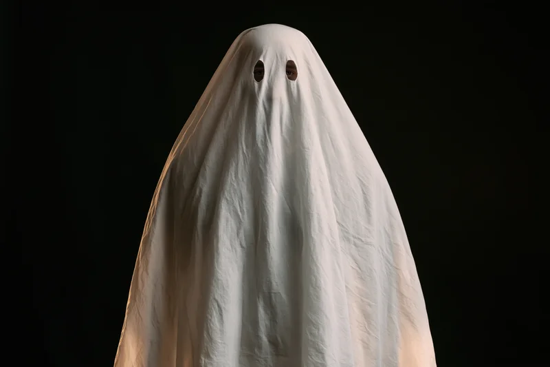 Person in white sheet ghost costume with eye holes against black background