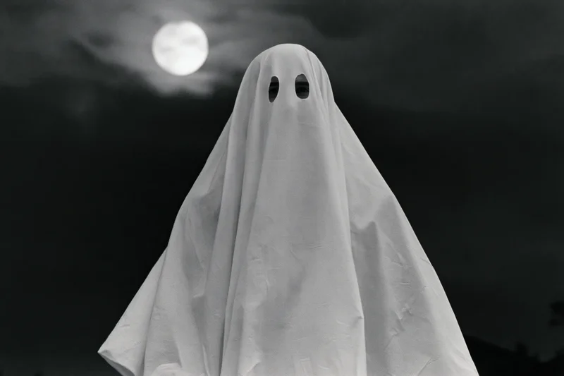 Ghost in white sheet costume with eye holes standing under full moon at night