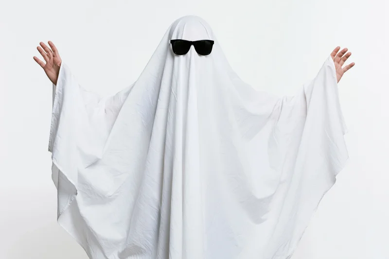 Person in white sheet ghost costume with sunglasses arms raised against white background