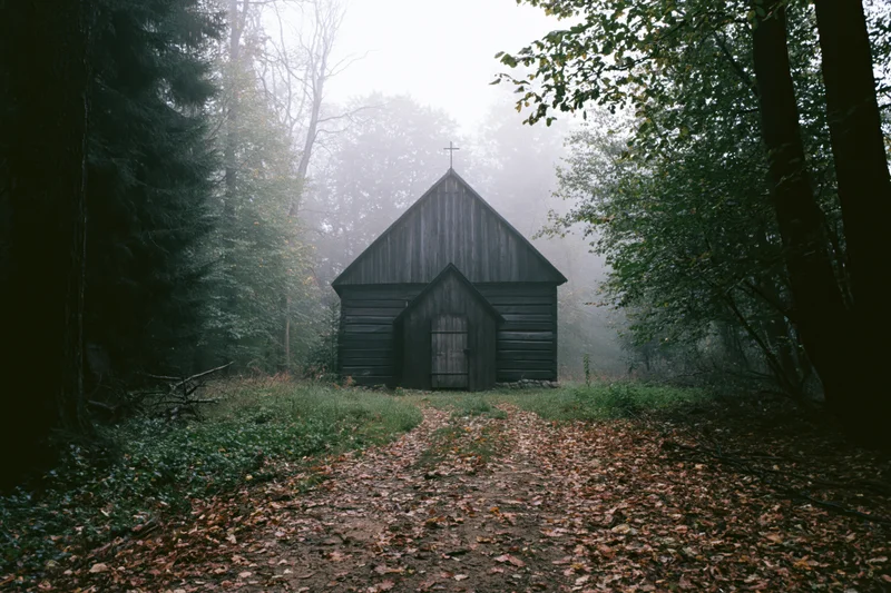 Small wooden chapel with cross on peaked roof in foggy forest, path leading through fallen leaves