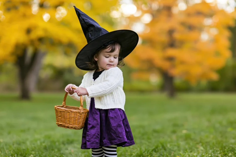Toddler girl in witch costume holding wicker basket in autumn park