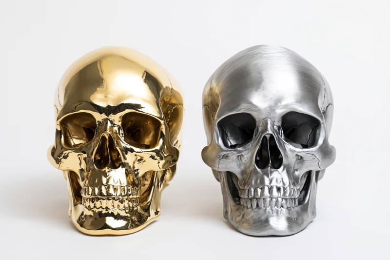 Two decorative human skull replicas in gold and silver side by side on white background