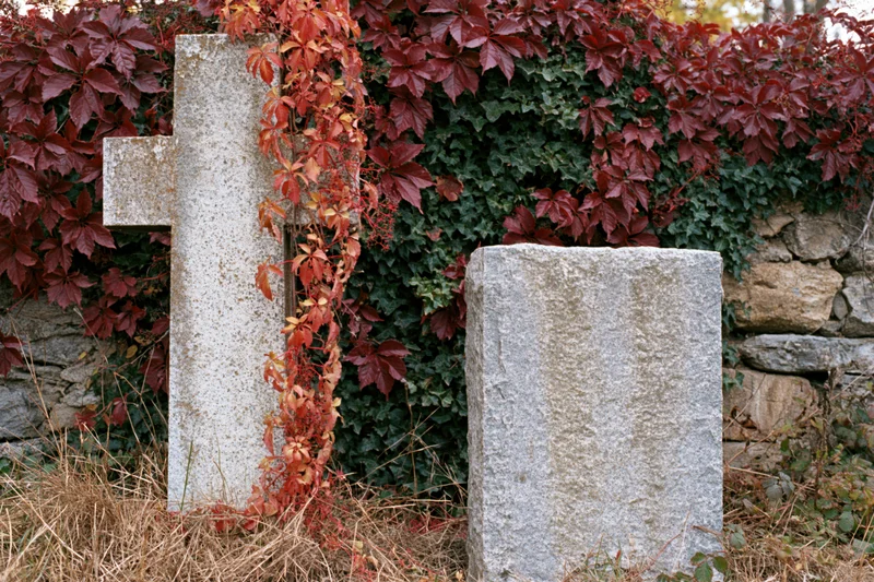 Two weathered granite grave markers in a small cemetery plot surrounded by autumn foliage and a stone wall
