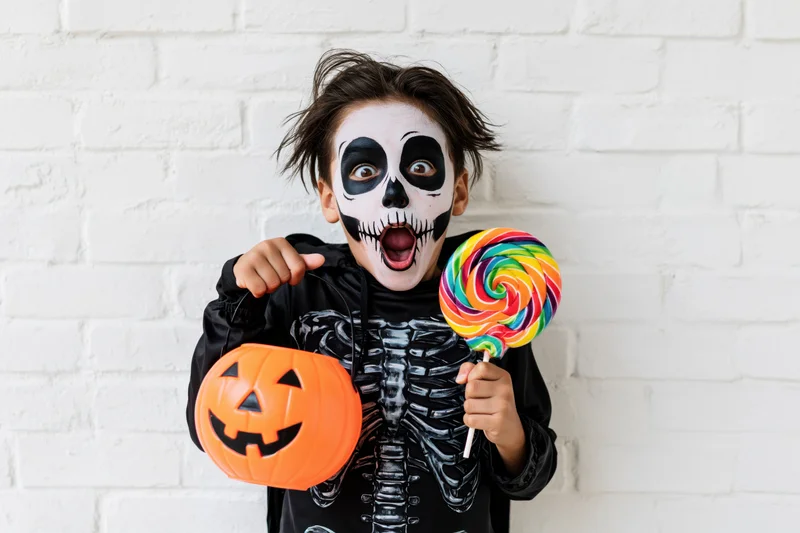 Boy in skeleton costume holding lollipop and pumpkin candy bag against white brick wall