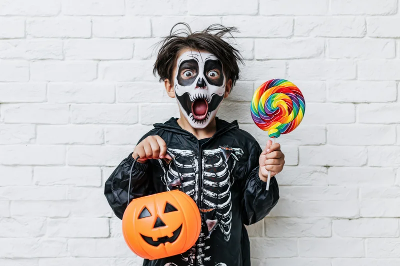 Boy in skeleton costume holding lollipop and candy bag against white brick wall