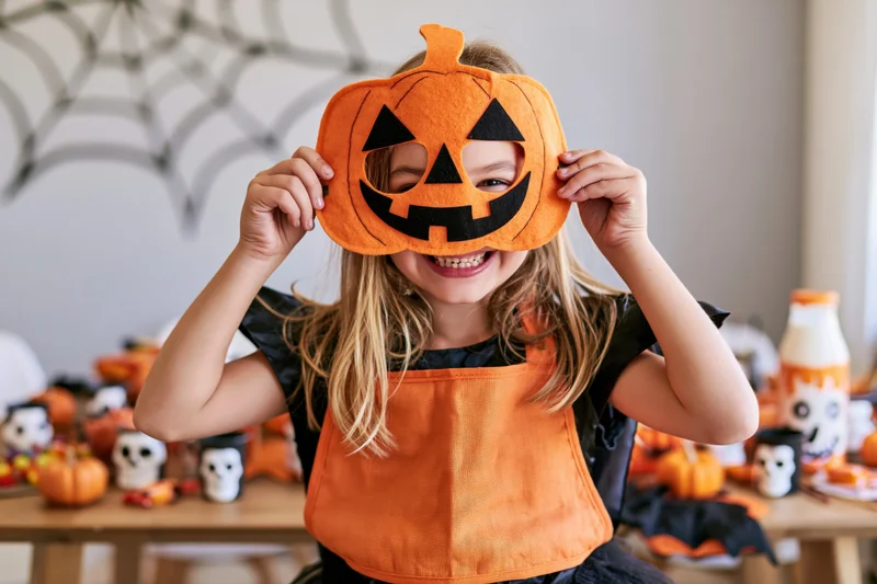 Young girl holding orange felt jack-o-lantern mask over her face at a halloween craft table