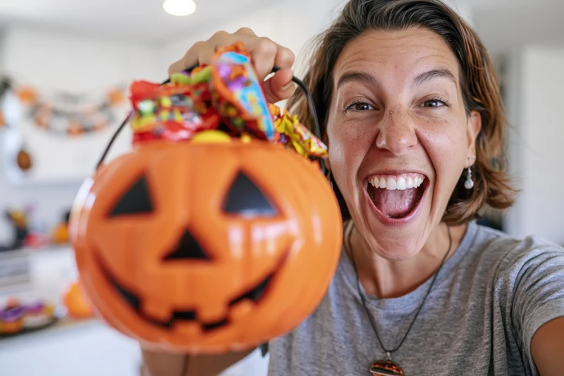Woman smiling widely while holding a plastic jack-o-lantern filled with candy indoors