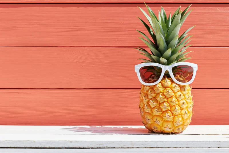 Pineapple wearing white sunglasses on a wooden deck against a coral wall