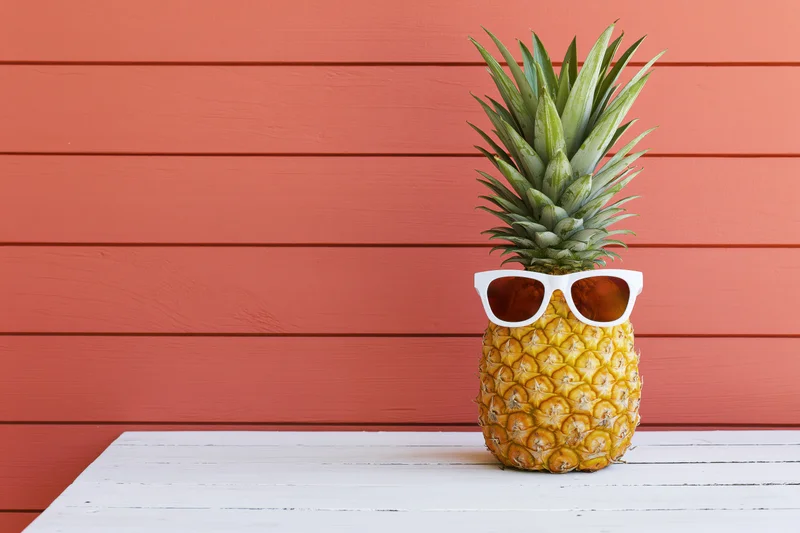 Whole pineapple wearing white-framed sunglasses on a white wooden table against orange wood paneling