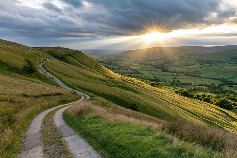 Winding path through rolling green hills at sunset with sun rays breaking through clouds over valley