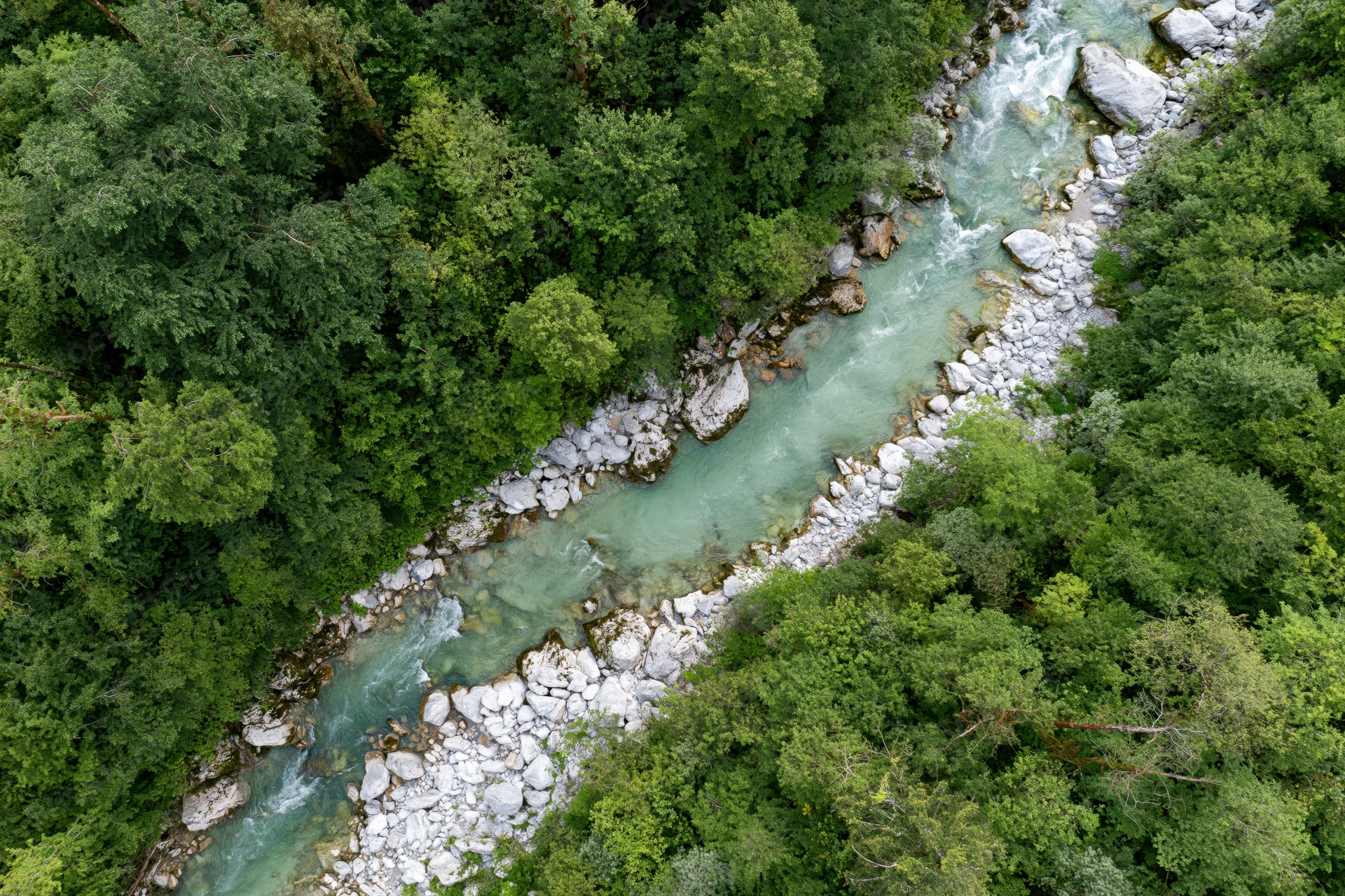Aerial top-down view of a narrow glacial river flowing through dense forested valley with rocks and boulders