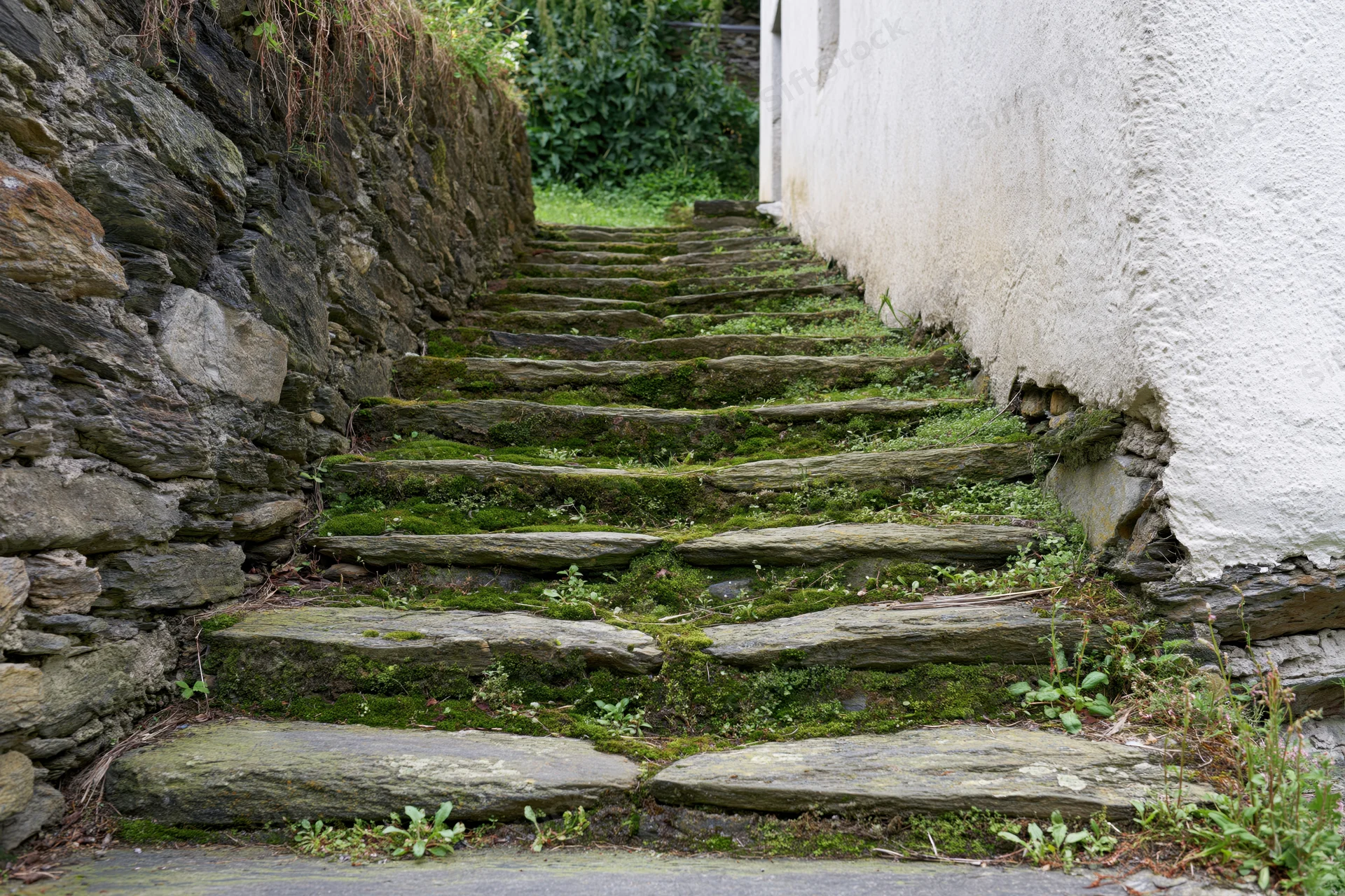 Moss-covered stone staircase ascending between a rough stone wall and a white plastered wall in a narrow outdoor passage