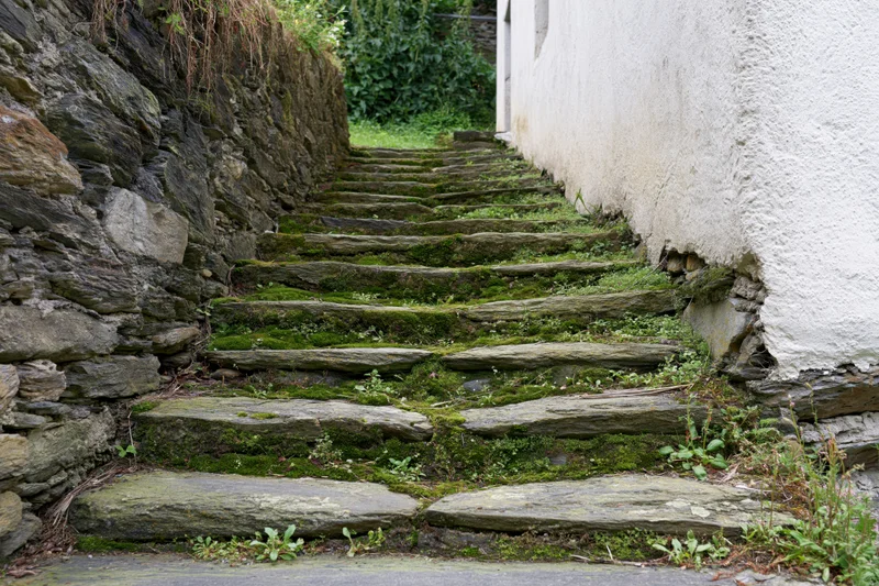 Moss-covered stone staircase ascending between a rough stone wall and a white plastered wall in a narrow outdoor passage