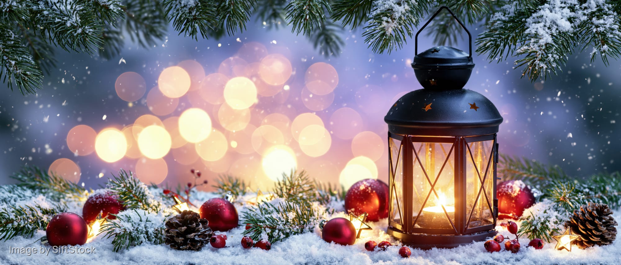 Black metal lantern with glowing candle on snowy ground surrounded by pine branches, red ornaments, and pinecones at night