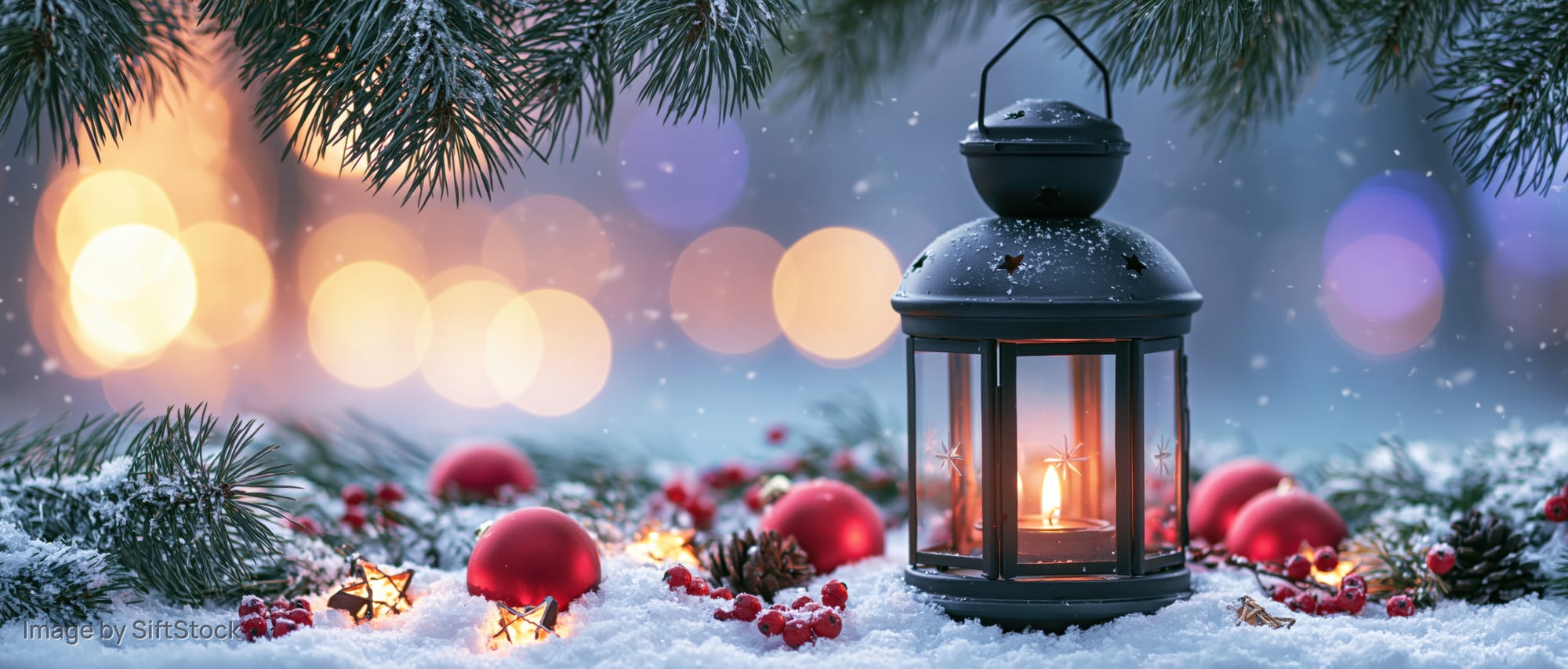 Black metal lantern with glowing candle on snowy ground surrounded by pine branches and red ornaments