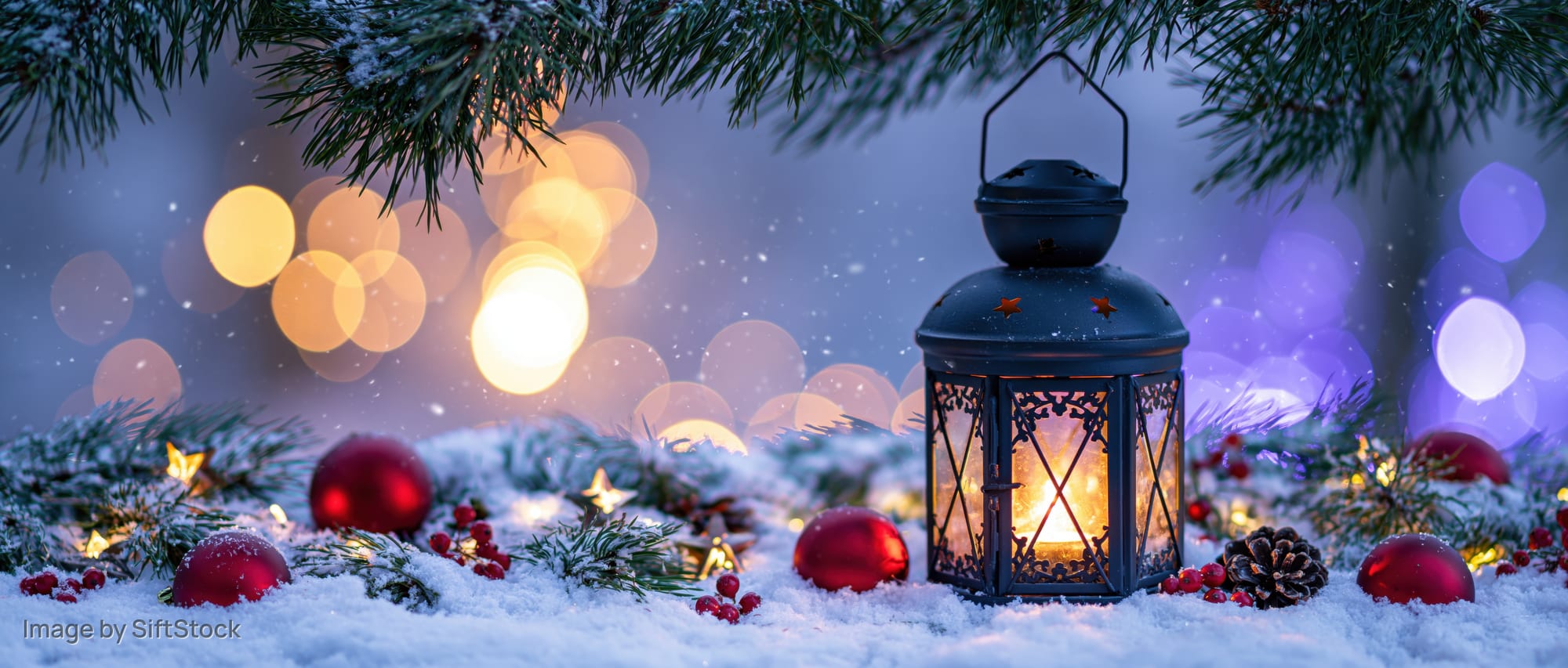 Black metal lantern with glowing candle on snowy ground surrounded by pine branches, red ornaments, and pinecones at night