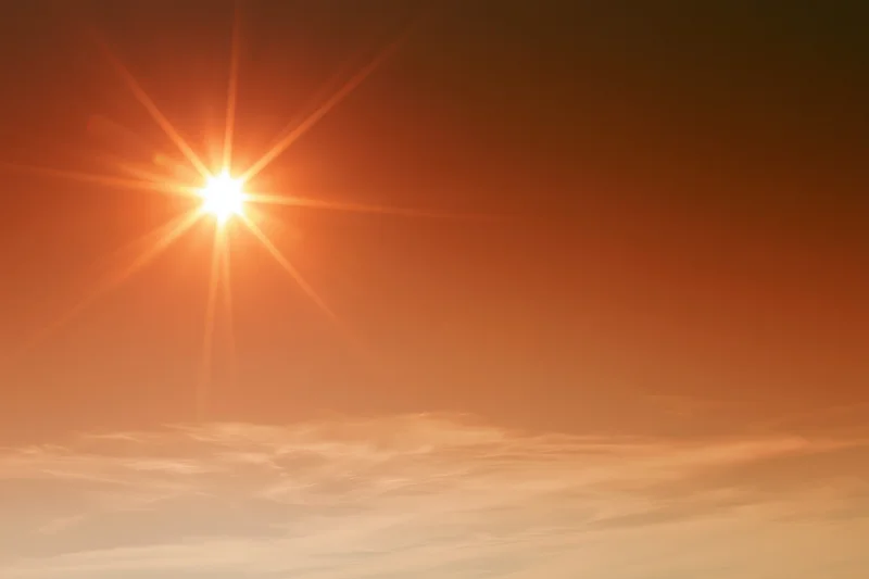 Blazing sun low in hazy summer sky viewed from below with lens flare and scattered clouds