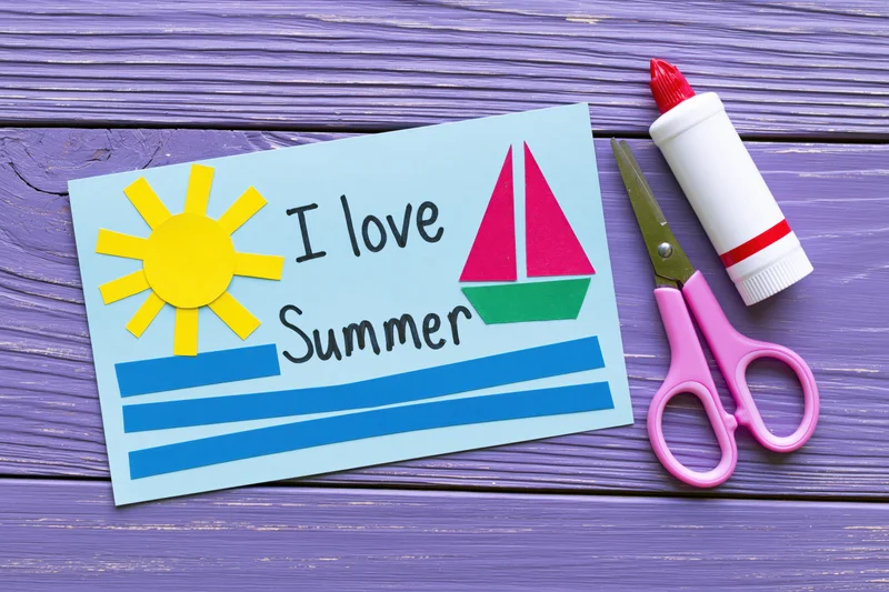 Handmade paper craft card with sun and sailboat reading I love Summer on purple wooden table with scissors and glue